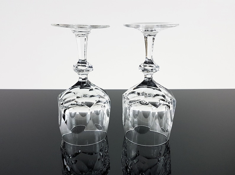 May include: Two clear crystal wine glasses positioned upside down. Each glass features a faceted, geometric bowl design and a multi-sectioned stem with a central hexagonal knob. The base of each glass is flat and circular, reflecting light clearly.