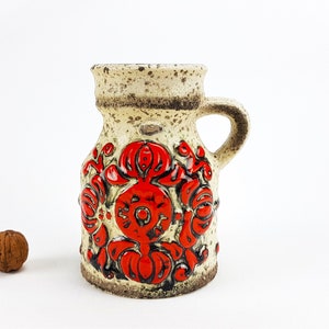 Könnte beinhalten: Ein kleiner, keramischer Krug mit Henkel. Der Krug ist mit einer weißen Glasur und einem roten und schwarzen Blumenmuster verziert.