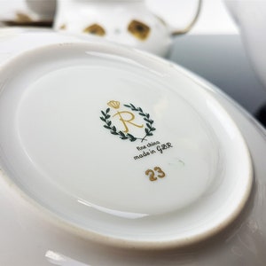May include: Close-up of a white porcelain saucer with a gold rim. The underside features a gold crown, a green wreath, the text "fine china made in GBR", and the number "23". Other white teacups and saucers are visible in the background.