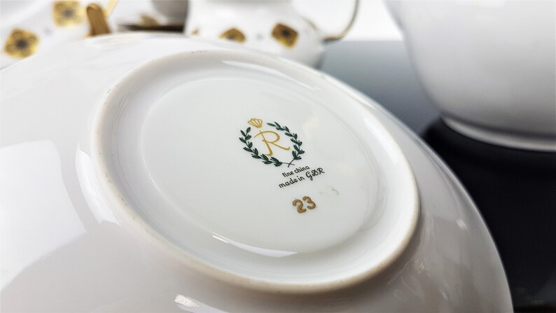 May include: Close-up of a white porcelain saucer with a gold rim. The underside features a gold crown, a green wreath, the text "fine china made in GBR", and the number "23". Other white teacups and saucers are visible in the background.