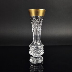 Könnte beinhalten: Eine klare Kristallvase mit Rautenmuster am Hals und am Fuß, getrennt durch einen glatten, vertikal gerippten Abschnitt. Der obere Rand ist mit einem breiten, massiven Goldband versehen, das einen deutlichen Kontrast zum Glaskörper bildet.
