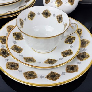 May include: A white teacup, saucer, and plate set with a gold rim and a repeating black and gold floral pattern. The teacup has a textured surface. The set is arranged on a reflective black surface, suggesting a formal dining setting.