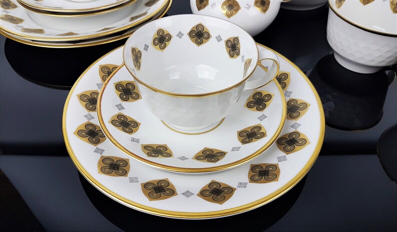 May include: A white teacup, saucer, and plate set with a gold rim and a repeating black and gold floral pattern. The teacup has a textured surface. The set is arranged on a reflective black surface, suggesting a formal dining setting.