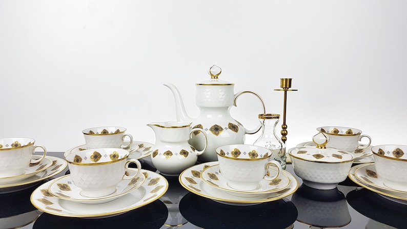 May include: A white porcelain tea set with gold trim and a brown floral pattern. The set includes teacups, saucers, a teapot, creamer, sugar bowl, and a coffee pot. A brass candlestick and small glass vase are also present.