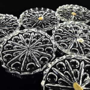 May include: A set of six circular clear glass coasters featuring a pressed pattern of radiating lines and small circular indentations. Each coaster has a scalloped edge and a small gold-colored sticker placed near the center of the glass surface.