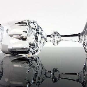 May include: Two clear crystal wine glasses featuring intricate geometric facets and a multi-sided stem design. One glass stands upright while the other rests horizontally, showcasing the reflective surfaces and the detailed cut glass patterns.