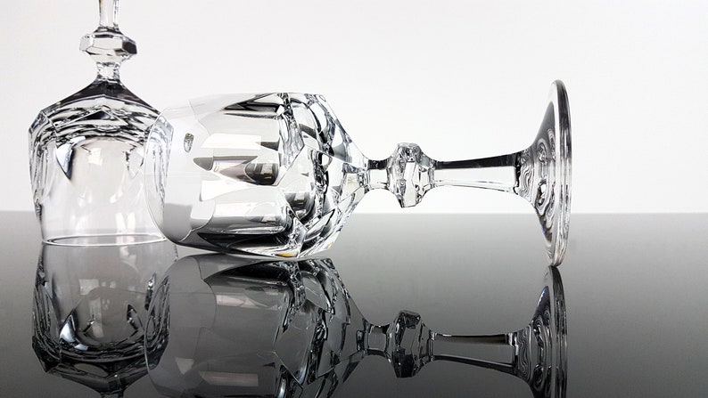 May include: Two clear crystal wine glasses featuring intricate geometric facets and a multi-sided stem design. One glass stands upright while the other rests horizontally, showcasing the reflective surfaces and the detailed cut glass patterns.