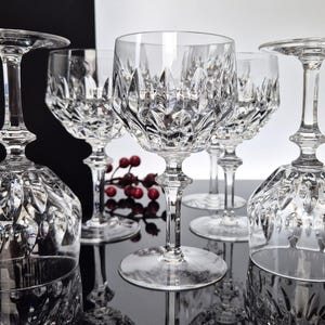 Könnte beinhalten: Eine Sammlung von Kristall-Weingläsern mit aufwendigen Diamantschliffmustern. Einige Gläser stehen aufrecht, andere sind umgedreht, um das detaillierte Design zu zeigen. Ein Zweig mit roten Beeren setzt einen Farbakzent vor einem schwarz-weißen Hintergrund.