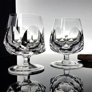 May include: Two clear glass brandy snifters with a diamond-shaped cut design. The snifters are on a reflective surface with a brown cigar.