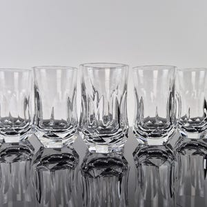 May include: Five clear glass tumblers with a faceted design. The glasses are arranged in a row on a reflective surface.