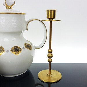 May include: A white porcelain tea set with gold trim and a brass candlestick. The tea set includes a teapot, cups, and saucers, decorated with black and gold floral patterns. The candlestick is tall with a round base and a cup for a candle.