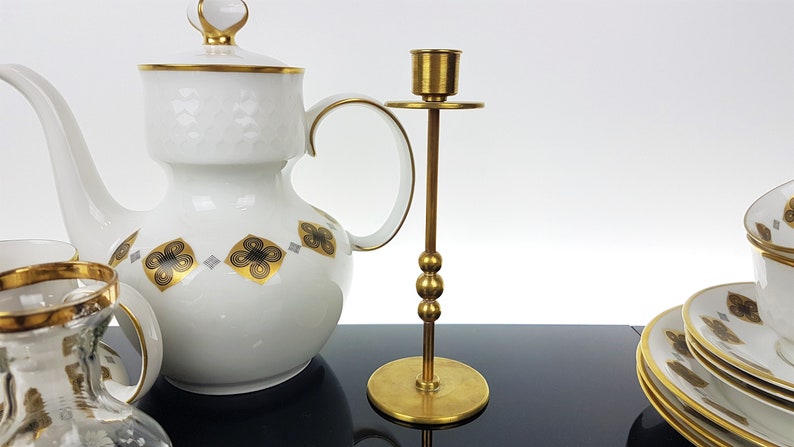 May include: A white porcelain tea set with gold trim and a brass candlestick. The tea set includes a teapot, cups, and saucers, decorated with black and gold floral patterns. The candlestick is tall with a round base and a cup for a candle.