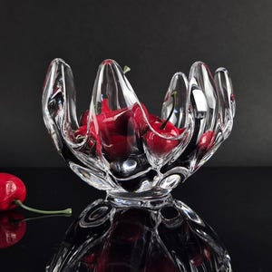 Puede incluir: Un cuenco de cristal transparente con un diseño de borde orgánico, ondulado y en forma de pétalo. El cuenco contiene varias cerezas de color rojo brillante con tallos verdes. Una cereza roja adicional con tallo verde se sitúa a la izquierda.