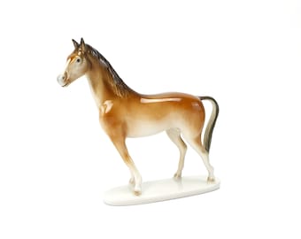 May include: A ceramic horse figurine in shades of brown and white. The horse is posed with its head turned, standing on a white oval base. The mane and tail are dark brown. The figurine has a glossy finish.