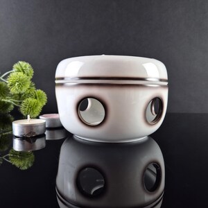 May include: A white ceramic essential oil diffuser with three circular openings and a brown trim. The diffuser is round and sits on a reflective surface. Small candles and green foliage are in the background.