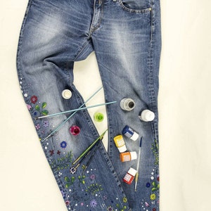 May include: A pair of blue denim jeans with a floral design painted on the legs. The jeans are laid out flat on a white surface with paint bottles and paintbrushes scattered around them.