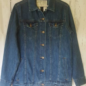 May include: A medium-wash blue denim jacket with a classic design. It features two chest pockets, side pockets, and a button-up front. The label reads "OVERSIZED DENIM JACKET". The jacket is hanging on a white hanger against a white wood background.