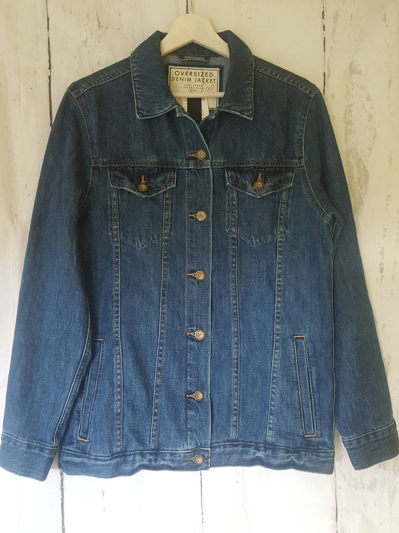 May include: A medium-wash blue denim jacket with a classic design. It features two chest pockets, side pockets, and a button-up front. The label reads "OVERSIZED DENIM JACKET". The jacket is hanging on a white hanger against a white wood background.