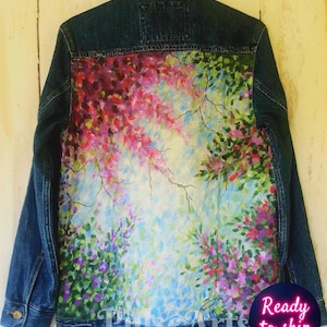 May include: A denim jacket with a vibrant, hand-painted floral design on the back. The artwork features a mix of pink, purple, green, and blue hues, creating a colorful and artistic statement piece. The jacket has a classic button-up design.