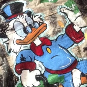 May include: A denim jacket with a hand-painted illustration of Scrooge McDuck, a cartoon character, wearing a top hat and bow tie. The background is a faded green and brown with dollar signs.