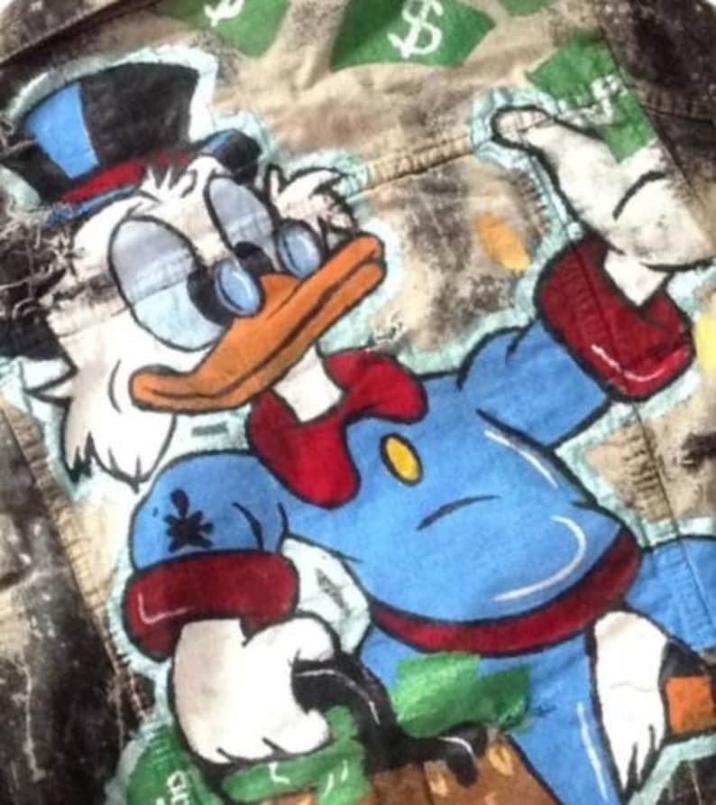 May include: A denim jacket with a hand-painted illustration of Scrooge McDuck, a cartoon character, wearing a top hat and bow tie. The background is a faded green and brown with dollar signs.