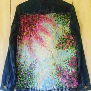 May include: A denim jacket with a vibrant, hand-painted floral design on the back. The artwork features pink, purple, and green blooms against a light blue and white background. The jacket has a classic button-down front and two chest pockets.