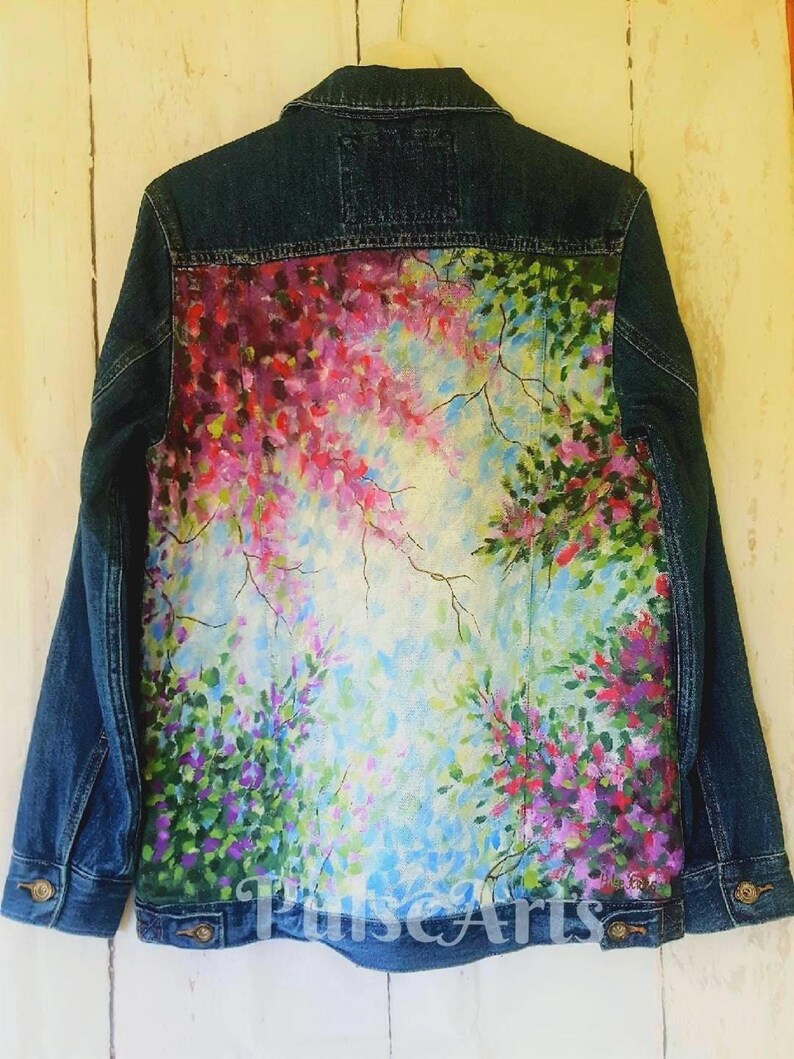 May include: A denim jacket with a vibrant, hand-painted floral design on the back. The artwork features pink, purple, and green blooms against a light blue and white background. The jacket has a classic button-down front and two chest pockets.