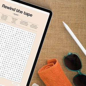 May include: A digital tablet with a word search puzzle on the screen. The puzzle is titled "Rewind the Tape - Music" and features a list of musicians and bands. The tablet is lying on a brown burlap surface with a rolled up orange towel and sunglasses.