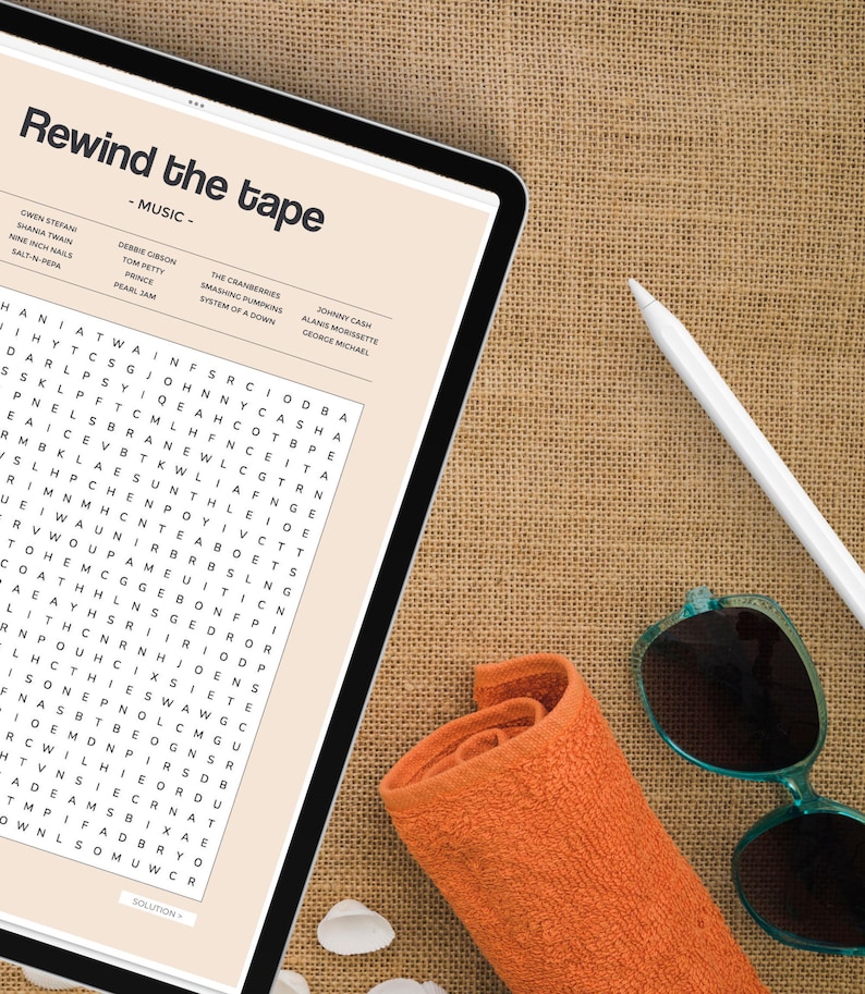 May include: A digital tablet with a word search puzzle on the screen. The puzzle is titled "Rewind the Tape - Music" and features a list of musicians and bands. The tablet is lying on a brown burlap surface with a rolled up orange towel and sunglasses.