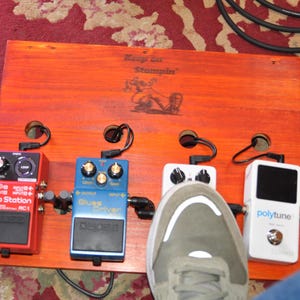 May include: A wooden pedalboard with four guitar effects pedals. The pedals are red, blue, white, and black. The pedalboard has the text "Keep on Stompin" and a graphic. A foot is on the pedalboard.