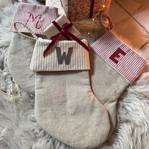 May include: Three beige linen Christmas stockings with striped cuffs. One stocking features a red embroidered M, another a dark grey embroidered W, and the third a red embroidered E. Each stocking has a hanging loop and a matching striped cuff.