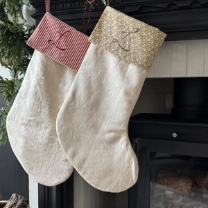 May include: Two cream-coloured fabric Christmas stockings hanging side by side. The left stocking features a red and white striped cuff with a stitched letter L. The right stocking has a tan floral patterned cuff with a stitched letter L.