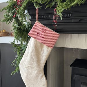 May include: A Christmas stocking featuring a plain off-white fabric body and a red and white striped cuff. The cuff is embroidered with a red cursive letter L. A matching red and white striped fabric loop is attached to the top for hanging purposes.