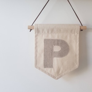 May include: A beige fabric pennant with a brown felt letter "P" hanging from a wooden dowel rod with brown leather straps.