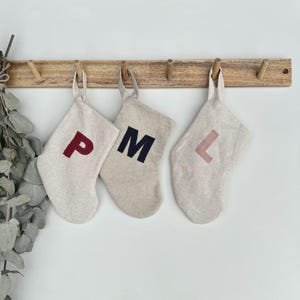 May include: Three beige linen stockings hanging from a wooden rack. Each stocking features a single capital letter in a different colour: a dark red P, a dark navy blue M, and a muted dusty pink L. The stockings have a simple, unlined design.