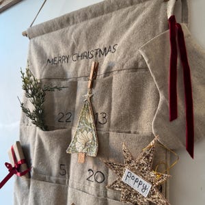 May include: A beige fabric advent calendar wall hanging with the text MERRY CHRISTMAS embroidered at the top. It features numbered pockets, a small fabric Christmas tree ornament, a glittery gold star ornament labelled poppy, and a stocking.