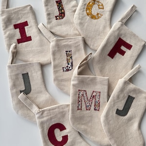 May include: A collection of ten off-white fabric stockings, each featuring a unique letter appliqué. The letters include various colours and patterns, such as solid red, dark grey, and floral prints, stitched onto the centre of each stocking.