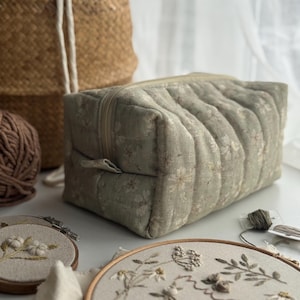 May include: A sage green quilted cosmetic bag with a floral pattern and a beige zip. The bag is rectangular and has a handle on the side. Embroidery hoops and yarn are in the background.