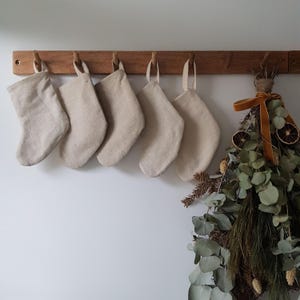 May include: Five small, plain beige fabric stockings with matching hanging loops are suspended from a horizontal wooden peg rack. Beside the stockings, a decorative bundle of dried eucalyptus, pine needles, and dried orange slices is tied with a ribbon.