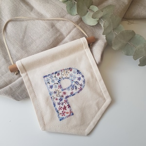 Peut inclure: Une bannière en toile blanche avec une lettre "P" en tissu à motif floral cousue dessus. La bannière a un bâtonnet en bois en haut et une ficelle pour l'accrocher.