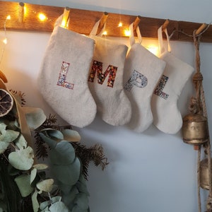 May include: Four beige Christmas stockings suspended from a wooden rail, each displaying a floral initial: L, M, P, and L. The stockings are lit by fairy lights, creating a warm, festive atmosphere. A dried floral arrangement is visible on the left.