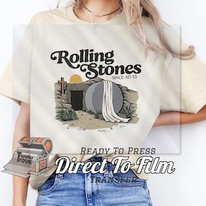 May include: Cream-colored t-shirt featuring a graphic of a tomb with a large stone, a cross, and the sun. The text "Rolling Stones Since AD 33" is above the graphic. Below the graphic is the text "READY TO PRESS Direct To Film TRANSFER".