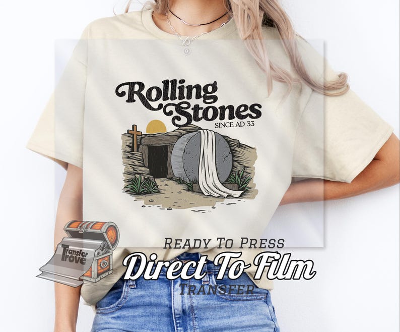 May include: Cream-colored t-shirt featuring a graphic of a tomb with a large stone, a cross, and the sun. The text "Rolling Stones Since AD 33" is above the graphic. Below the graphic is the text "READY TO PRESS Direct To Film TRANSFER".