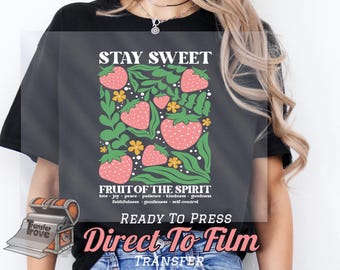 May include: A black t-shirt with a graphic design of strawberries and flowers. The text "Stay Sweet" is at the top of the design, and the text "Fruit of the Spirit" is below the design. The text "love, joy, peace, patience, kindness, goodness, faithfulness, gentleness, self-control" is below the text "Fruit of the Spirit". The text "Ready to Press Direct To Film Transfer" is at the bottom of the design.
