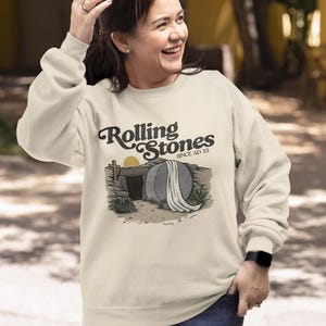 May include: Cream-colored sweatshirt with the text "Rolling Stones Since AD 33" above an illustration of a tomb with a cross and a rising sun. The sweatshirt is long-sleeved and has a crew neck.