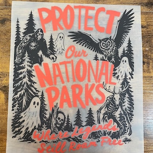 May include: A rectangular transfer sheet featuring the red text PROTECT Our NATIONAL PARKS Where Legends Still Roam Free. The design includes black illustrations of a sasquatch, an owl, ghosts, a skeleton creature, and pine trees.