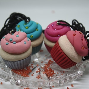 May include: Four miniature cupcake charms with colourful frosting and sprinkles. The cupcakes are on a clear glass plate with red sugar crystals scattered around.