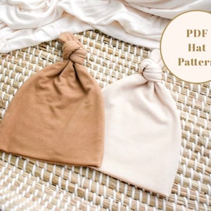 Puede incluir: Dos gorros de punto con un nudo en la parte superior, uno es marr&oacute;n y el otro beige. Los gorros est&aacute;n sobre una estera tejida con un fondo blanco. El texto "PDF Hat Pattern" est&aacute; en un c&iacute;rculo en la parte superior derecha de la imagen.