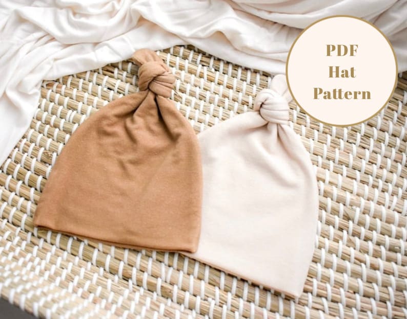 Puede incluir: Dos gorros de punto con un nudo en la parte superior, uno es marr&oacute;n y el otro beige. Los gorros est&aacute;n sobre una estera tejida con un fondo blanco. El texto "PDF Hat Pattern" est&aacute; en un c&iacute;rculo en la parte superior derecha de la imagen.
