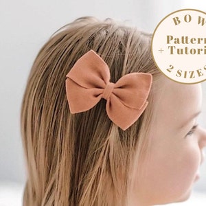 May include: A close-up of a young girl wearing a peach-colored fabric bow in her hair. The bow is tied with a knot in the center and has two loops on either side. The image includes text that reads "BOW Pattern + Tutorial 2 SIZES".